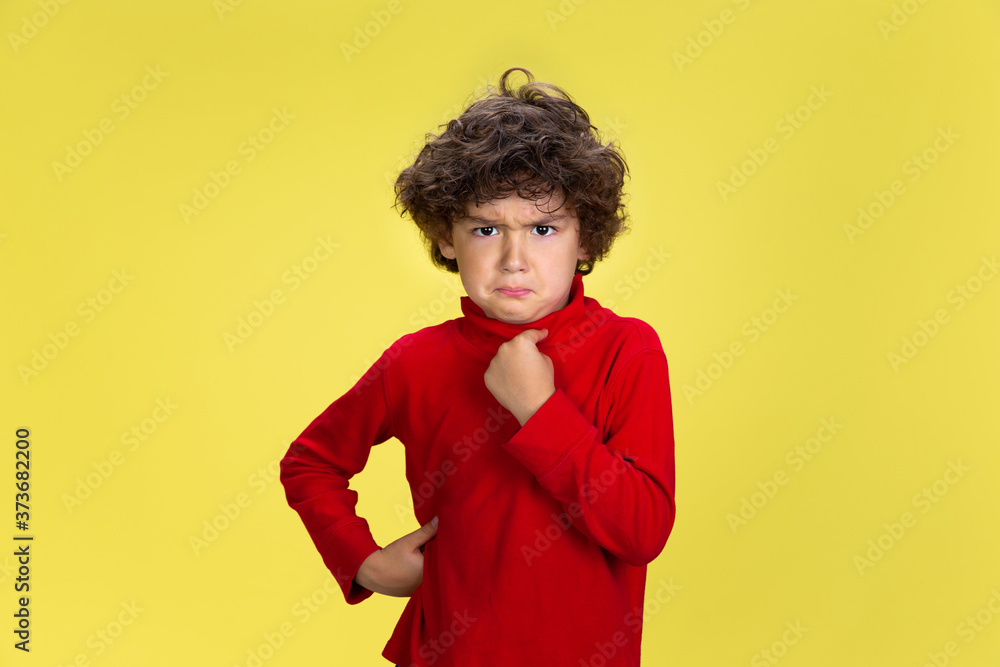 Pointing at himself. Portrait of pretty young curly boy in red wear on yellow studio background. Childhood, expression, education, fun concept. Preschooler with bright facial expression and sincere