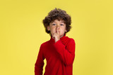 Whispering. Portrait of pretty young curly boy in red wear on yellow studio background. Childhood, expression, education, fun concept. Preschooler with bright facial expression and sincere emotions.
