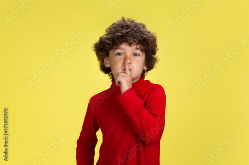 Whispering. Portrait of pretty young curly boy in red wear on yellow studio background. Childhood, expression, education, fun concept. Preschooler with bright facial expression and sincere emotions.
