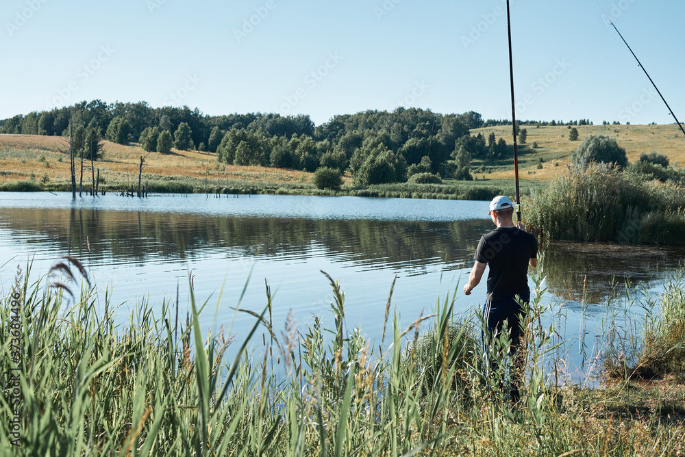 Summer fishing for fishing rod. A fisherman stands against a background of a beautiful, calm lake with reflection of trees. A beautiful scene of outdoor activities Summer fishing for fishing rod. A fisherman stands against a background of a beautiful, calm lake with reflection of trees. A beautiful scene of outdoor activities