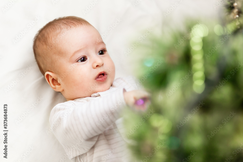 Baby boy or girl lying under Christmas tree. Little child plays with fir tree decorated for New Year celebration. Cute kid portrait. Winter holiday spirit. Baby boy or girl lying under Christmas tree. Little child plays with fir tree decorated for New Year celebration. Cute kid portrait. Winter holiday spirit.