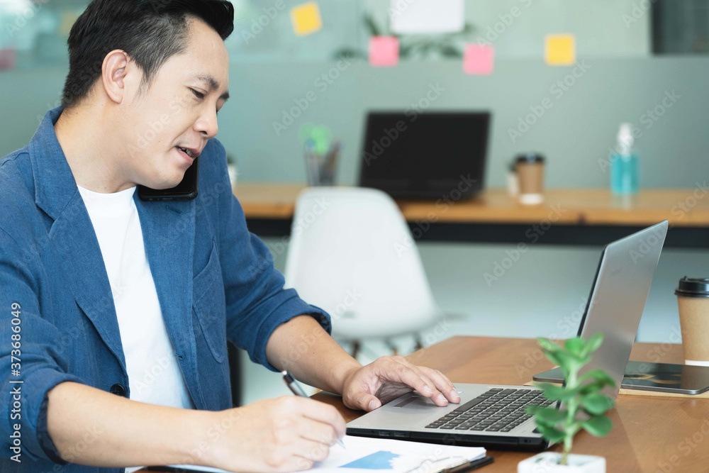 Handsome asian businessman working with laptop in office,watching on laptop holding call talking on the phone with clients partners and writing down ideas in a notebook.Busy concept,Selective focus Handsome asian businessman working with laptop in office,watching on laptop holding call talking on the phone with clients partners and writing down ideas in a notebook.Busy concept,Selective focus
