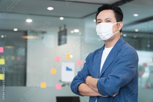 Young smart asian business man wearing medical face mask looking and standing with arms folded in modern office under quarantine from coronavirus or COVID-19,copy space