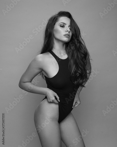 Sexy tanned caucasian woman with long brunette wavy hair and perfect natural makeup in black body or swimswuit posing against azure wall. Fashion studio portrait