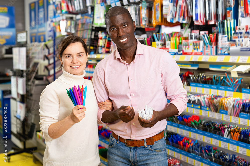 Cheerful positive smiling African man and European woman choosing pens and pencils in office supply shop