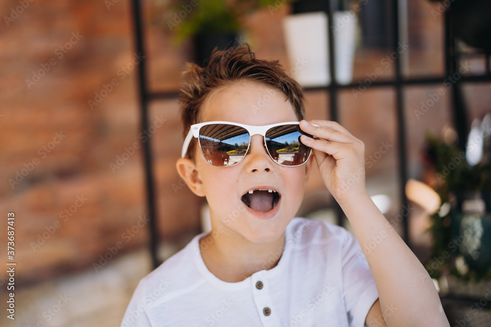 cute cauasian boy in white tee shirt and sunglasses posing for camera and showing his lost tooth. brickwall and black shelving unit on background. Image with selective focus