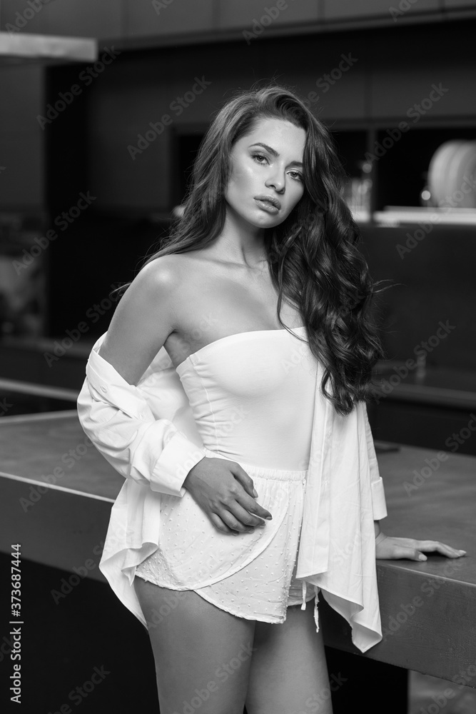 Fototapeta premium Young beautiful caucasian woman with perfect natural makeup and wavy brunette hair wearing white body, shorts and jacket standing and posing at dark stylish kitchen