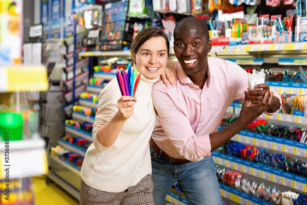 Portrait of positive smiling Afro American man and Caucasian woman choosing ball pens at stationery store