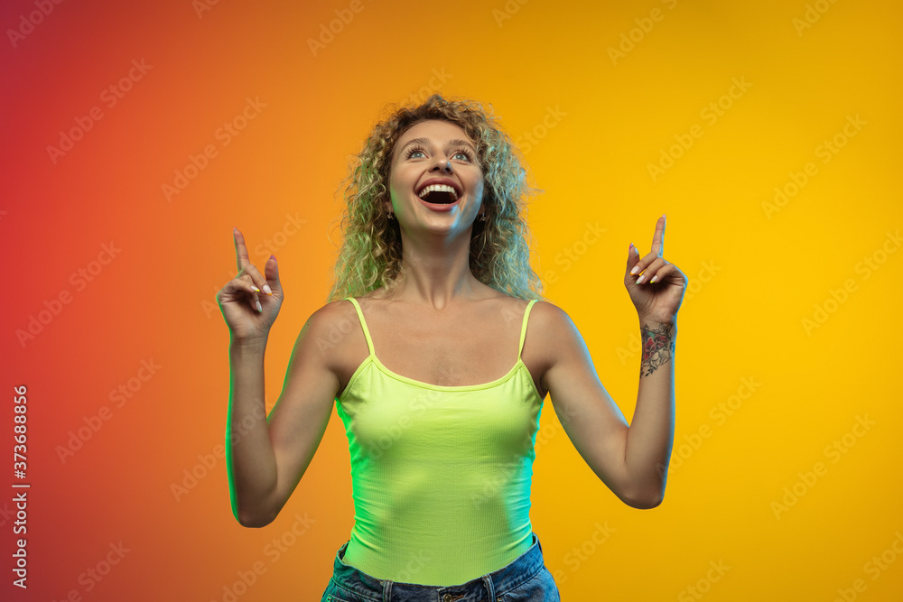 Pointing. Caucasian young woman's portrait isolated on gradient studio background in neon. Beautiful female curly model in casual style. Concept of human emotions, facial expression, youth, sales, ad. Pointing. Caucasian young woman's portrait isolated on gradient studio background in neon. Beautiful female curly model in casual style. Concept of human emotions, facial expression, youth, sales, ad.