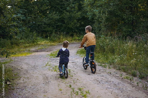 back view of cute caucasian boys brothers are cycling together in countryside in summer time. Little one is riding runbike, elder one standing on pedals of bicycle. Image with selective focus