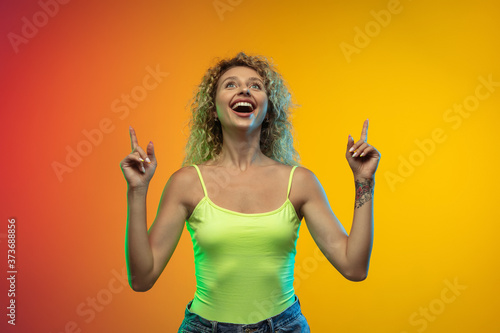 Pointing. Caucasian young woman's portrait isolated on gradient studio background in neon. Beautiful female curly model in casual style. Concept of human emotions, facial expression, youth, sales, ad.