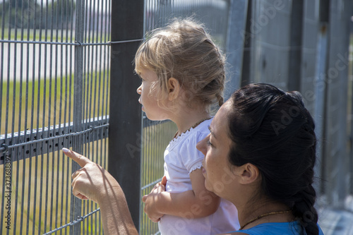 Photography A woman and a child look into the distance through a fence with a small net, the mother points at something to the child