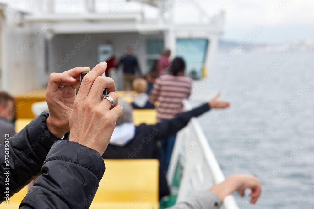 Hands of a man holding a smartphone and taking a photo during the ferry trip. Fuzzy silhouettes of other people in the background. Hands of a man holding a smartphone and taking a photo during the ferry trip. Fuzzy silhouettes of other people in the background.