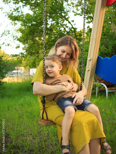 Cute baby boy and mother ride on swing in playground on green lawn. Summertime photo for ad or blog about motherhood © Georgii