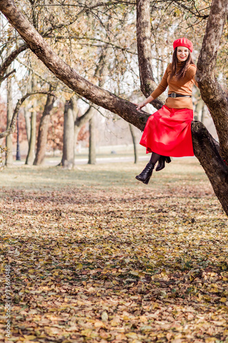 Beautiful brunette woman in a red beret, brown turtleneck and red skirt sits high on a tree branch in an autumn park