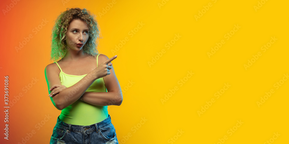 Pointing. Caucasian young woman's portrait on gradient studio background in neon. Beautiful female curly model in casual style. Concept of human emotions, facial expression, youth, sales, ad. Flyer. Pointing. Caucasian young woman's portrait on gradient studio background in neon. Beautiful female curly model in casual style. Concept of human emotions, facial expression, youth, sales, ad. Flyer.