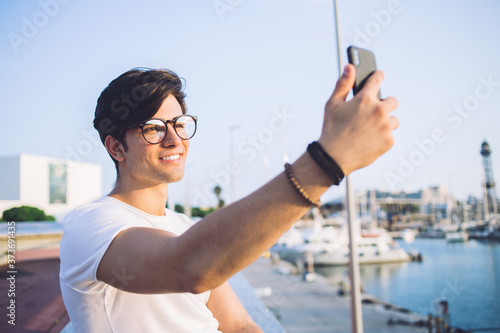 Smiling caucasian male travel blogger holding mobile phone making selfie in harbor on sunny day, positive prosperous man influencer making picture on smartphone sitting in marine port on weekends