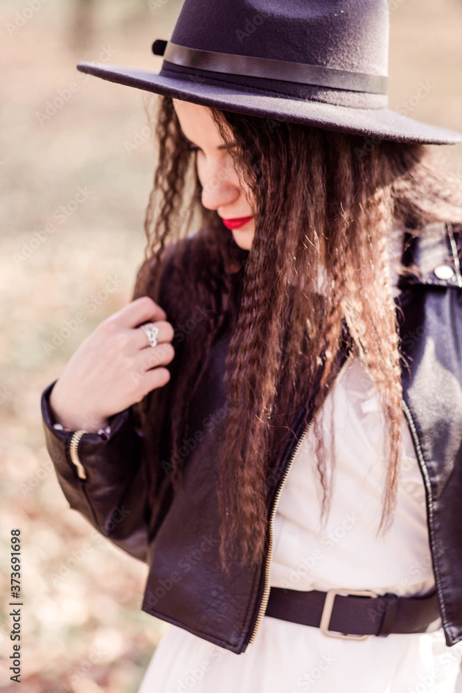 Beautiful brunette girl with long hair in a white dress, black leather jacket and black hat posing in an autumn park Beautiful brunette girl with long hair in a white dress, black leather jacket and black hat posing in an autumn park