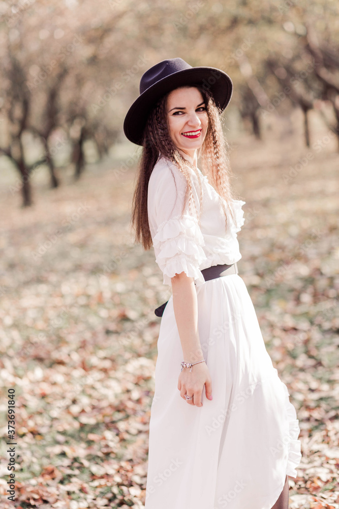 Beautiful brunette girl with long hair in a white dress and black hat posing in an autumn park. Girl looking at the camera and smiling
