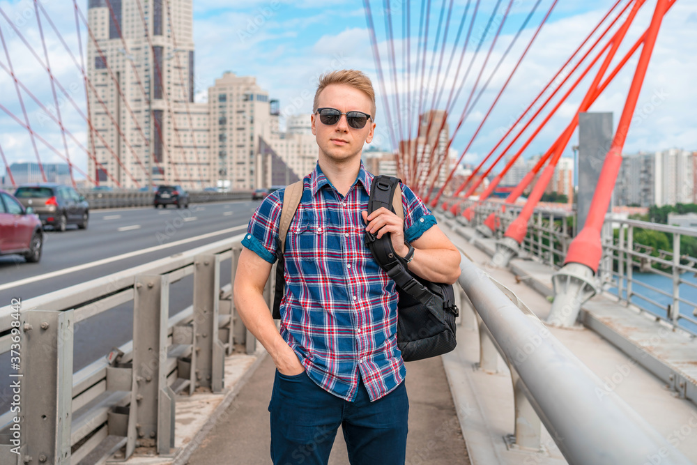 A young man in sunglasses stands on the picturesque red bridge in Moscow, car traffic nearby in the photo