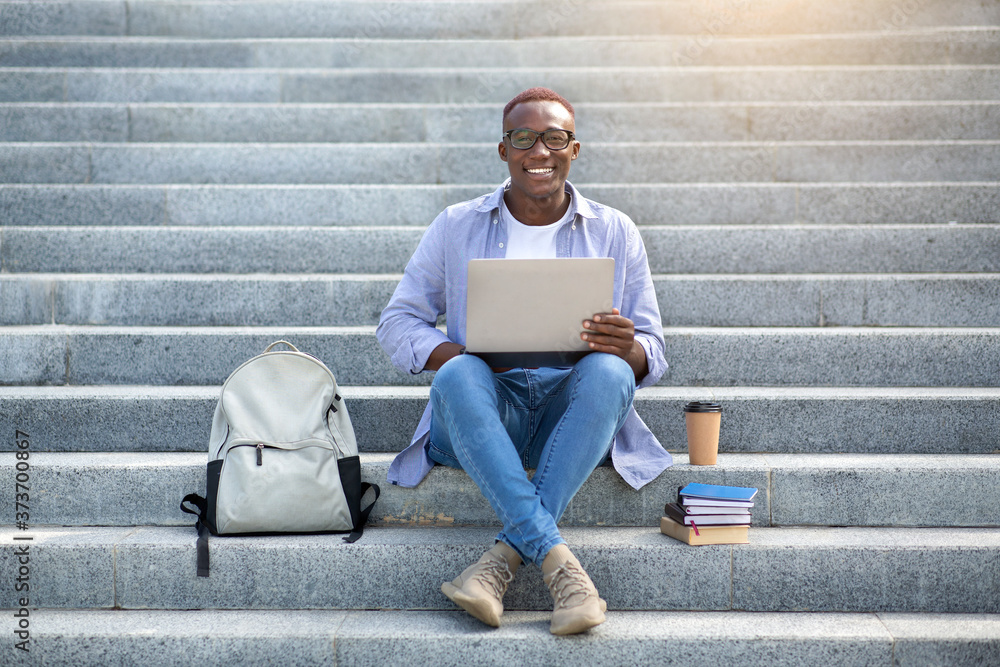 Online education. Intelligent black man with laptop and pile of books sitting on stairs downtown