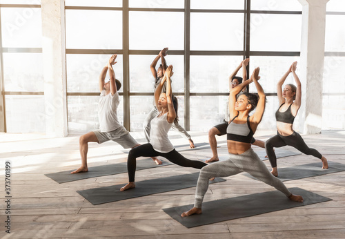 Group Of Young Yoga Enthusiasts Training Together In Studio, Practicing Warrior Pose