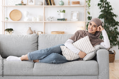 Relaxed arabic woman lying on couch at home, cuddling pillow, enjoying weekend