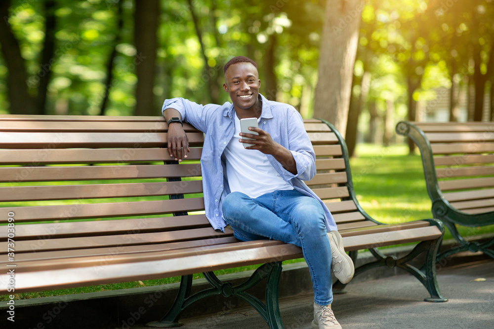 Smiling black man with mobile phone relaxing on bench at park on summer day Smiling black man with mobile phone relaxing on bench at park on summer day