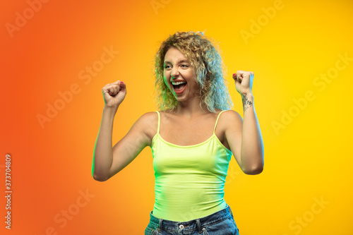 Celebrating. Caucasian young woman's portrait isolated on gradient studio background in neon. Beautiful female curly model in casual style. Concept of human emotions, facial expression, youth, sales