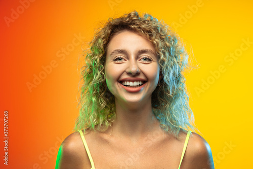 Smiling. Close up caucasian young woman's portrait on gradient studio background in neon. Beautiful female curly model in casual style. Concept of human emotions, facial expression, youth, sales, ad.
