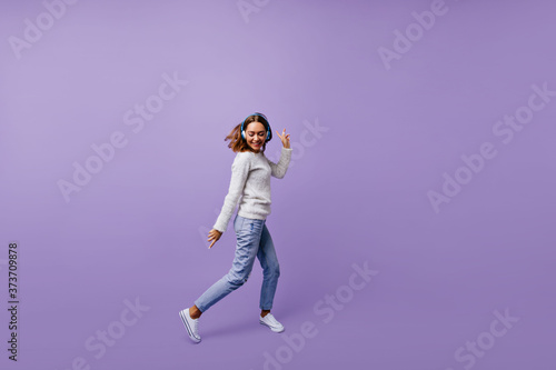 Dancing Slavic female student in stylish jeans and winter sweater moves on purple background. Brunette with headphones poses for full-length photo