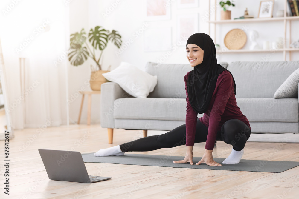 Sporty muslim woman in hijab exercising at home, watching tutorials on laptop Sporty muslim woman in hijab exercising at home, watching tutorials on laptop