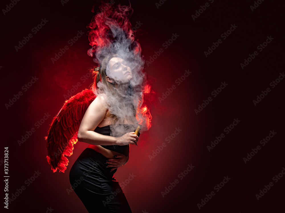 Girl with a VAPE on a red background. A woman in a cloud of smoke from an electronic cigarette. The girl poses and smokes a VAPE. A Vaper with red wings. Vaping fantasy.