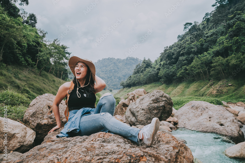 Happy Traveler Asian woman with backpack laughing while sitting on the rock by river in mountain during vacation. Travel in forest concept