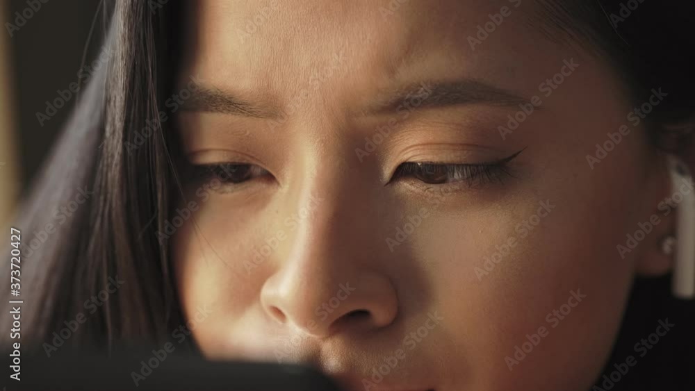 A close-up view of a concentrated young asian woman with earbuds is sitting on the sofa and watching something in her smartphone at home A close-up view of a concentrated young asian woman with earbuds is sitting on the sofa and watching something in her smartphone at home