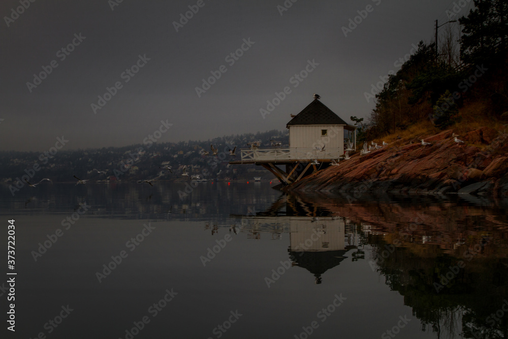 A white bathing house reflects in the calm surface of the fjord