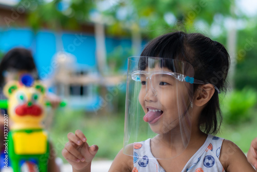 An Asian girl aged 3 - 4 wearing a face shield is playing in front of her house.