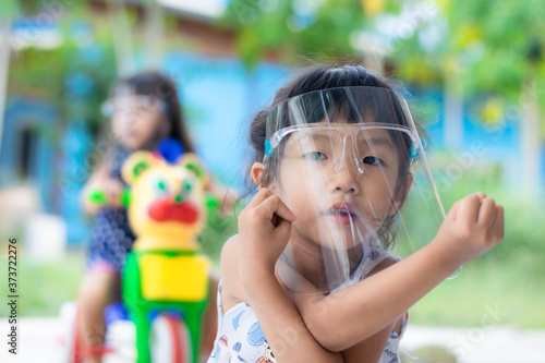 An Asian girl aged 3 - 4 wearing a face shield is playing in front of her house.