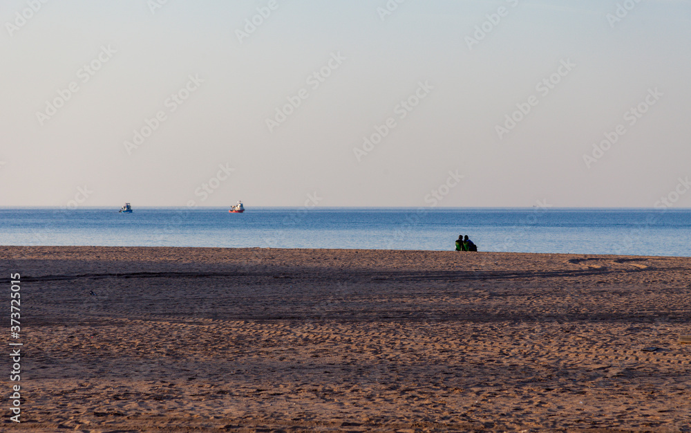 Couple sitting on the beach and watches the fishing boats pass by. Couple alone in the beach near the sea and watching the ocean. Fishing boat is in focus. Romance at Sunset time.