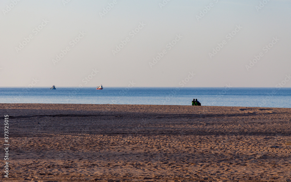 Couple sitting on the beach and watches the fishing boats pass by. Couple alone in the beach near the sea and watching the ocean. Fishing boat is in focus. Romance at Sunset time. Couple sitting on the beach and watches the fishing boats pass by. Couple alone in the beach near the sea and watching the ocean. Fishing boat is in focus. Romance at Sunset time.