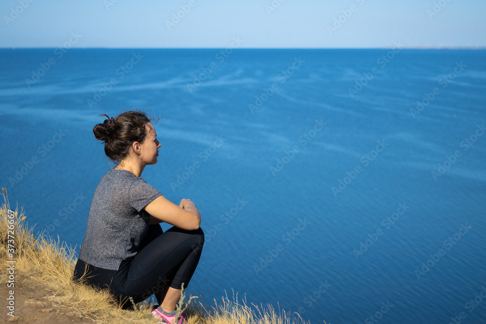 Side view of young brunette woman sitting at edge of mountain cliff looking at blue sea, meditating relaxing alone, enjoying beautiful nature scenery landscape in sunny summer day. Active lifestyle