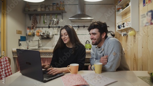 Smiling couple drinking coffee at kitchen. Joyful couple watching laptop computer. Happy man talking with woman at kitchen.
