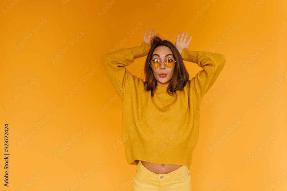Positive tanned girl in sunglasses making funny faces on orange background. Indoor portrait of cheerful brown-haired woman wears orange sweater. Positive tanned girl in sunglasses making funny faces on orange background. Indoor portrait of cheerful brown-haired woman wears orange sweater.