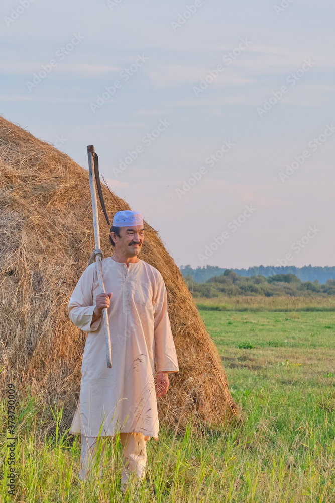 Φωτογραφία An senior Muslim man in a skullcap and traditional clothes leaves with a hand scythe from the hay field at sunset