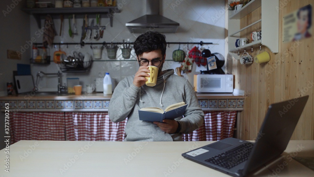 Young man reading a book and drinking tea in kitchen at home at night Young man reading a book and drinking tea in kitchen at home at night