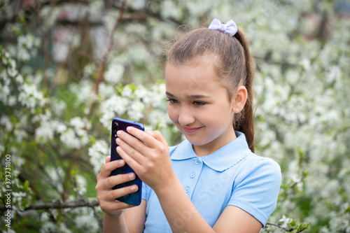 Little Girl Admires Smartphone On Background Of Blooming Garden In Spring Close-Up.