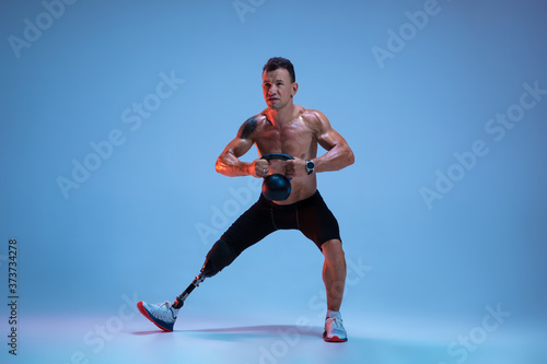 Athlete with disabilities or amputee isolated on blue studio background. Professional male sportsman with leg prosthesis training with weights in neon. Disabled sport and overcoming  wellness concept.