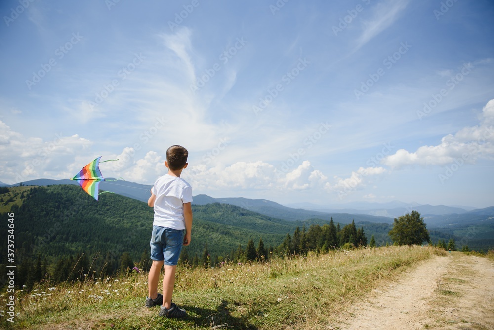 Little school age boy running down the slope with kite in the sun. Sunny summer or spring day at sunset. Active outdoor games and leisure. Mountain landscape with hills and coniferous trees.
