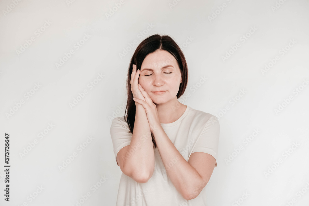 Beautiful brunette woman with eyes closed keeps hands on cheek, dressed in casual white t shirt, has minimal makeup, poses indoor on a white background. Copy, empty space for text Beautiful brunette woman with eyes closed keeps hands on cheek, dressed in casual white t shirt, has minimal makeup, poses indoor on a white background. Copy, empty space for text