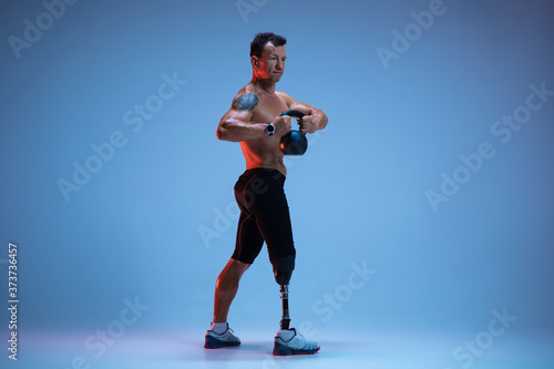 Fototapeta Naklejka Na Ścianę i Meble -  Athlete with disabilities or amputee isolated on blue studio background. Professional male sportsman with leg prosthesis training with weights in neon. Disabled sport and overcoming, wellness concept.